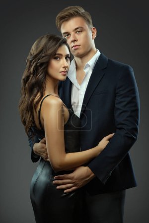 Téléchargez les photos : Couple de mode sur Gray. Homme élégant en costume embrassant belle fille en robe noire de cocktail. Femme romantique avec beau petit ami datant - en image libre de droit