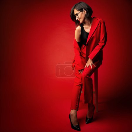 Téléchargez les photos : Femme de mode en costume rouge. Modèle élégant avec Bob noir coupe de cheveux dans les lunettes de vue en costume pantalon assis sur fond rouge studio montrant les jambes - en image libre de droit