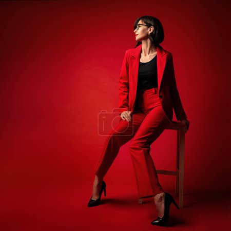 Téléchargez les photos : Femme d'affaires Mode. Elégante Lady en costume de pantalon rouge avec Bob Coiffure portant des lunettes de vue de côté noir. Modèle élégant dans les vêtements de luxe - en image libre de droit