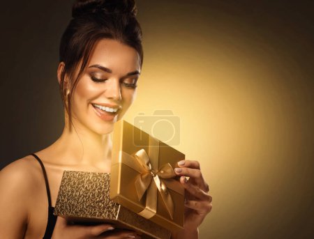 Téléchargez les photos : Cadeau pour Femme. Belle fille ouverture cadeau boîte avec la lumière brillante. Joyeux sourire Modèle recevoir Surprise sur fond doré - en image libre de droit