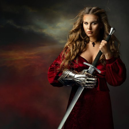 Téléchargez les photos : Belle guerrière à l'épée. Chevalier Féminin Médiéval en Robe Rouge avec Gant Armure. Belle fille viking en robe de fantaisie sur fond de ciel sombre - en image libre de droit