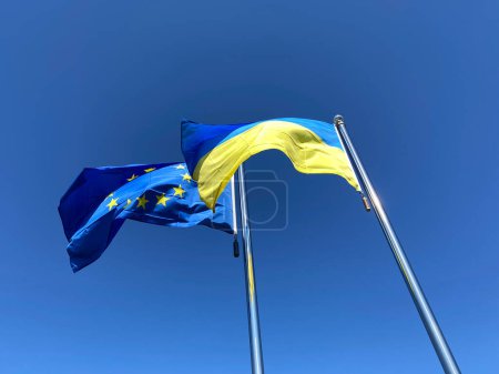 Photo for Ukrainian and European Union flags waved on flagpoles against blue sky - Royalty Free Image