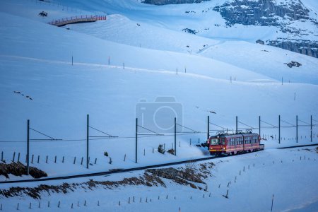 Téléchargez les photos : Un train à crémaillère circule sur le chemin de fer de Jungfraujoch (haut de l'Europe) à Kleine Scheidegg sur la colline enneigée avec Eiger, Monch et Jungfrau en arrière-plan dans les hautes terres bernoises, en Suisse - en image libre de droit
