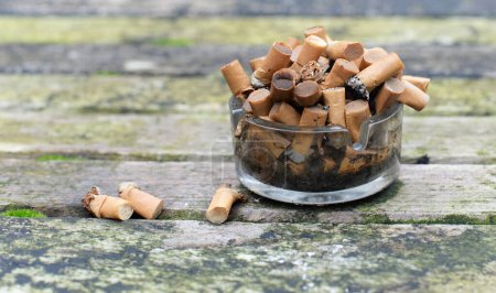 Photo for Ashtray full of soaked cigarette buds and ashes on weathered teak wooden table - Royalty Free Image