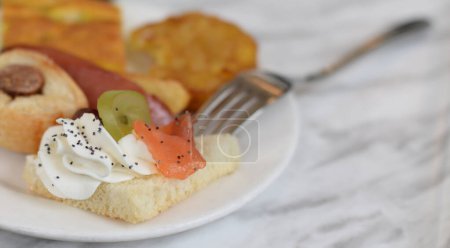 Toast mit Frischkäse und Räucherlachs bestreut mit Mohn auf Teller mit italienischen Vorspeisen auf Marmortisch, selektiver Fokus