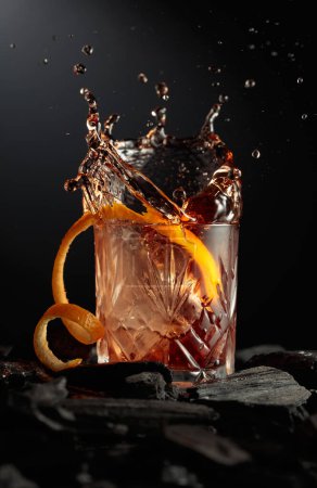 Téléchargez les photos : Un morceau de glace tombe dans le verre avec un cocktail. Cocktail avec glace et écorce d'orange sur fond sombre. - en image libre de droit