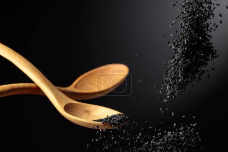 Téléchargez les photos : Les grains de sésame noir sont versés avec des cuillères en bois. Sésame noir sur fond sombre. Espace de copie. - en image libre de droit
