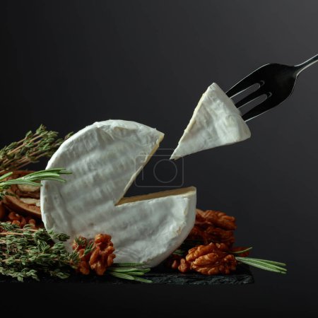 Brie cheese with walnuts, thyme, and rosemary on a black background. Brie cheese and fork with triangle piece.