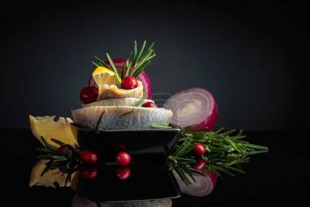 Photo for Herring fillet in oil with lemon, red onion, and rosemary garnished with cranberries. Black dish with herring on a black reflective background. - Royalty Free Image
