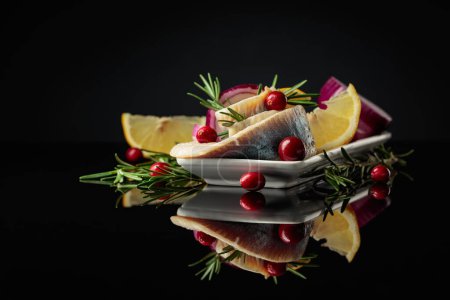 Photo for Herring fillet in oil with lemon, red onion, and rosemary garnished with cranberries. White dish with herring on a black reflective background. - Royalty Free Image