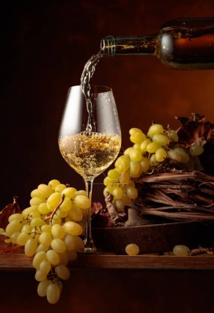 Foto de El vino blanco se vierte en una copa. Vino blanco y racimo de uvas en mesa de madera vintage. - Imagen libre de derechos