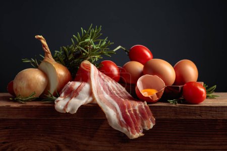 Foto de Panceta ahumada con huevos, tomates, cebollas y romero en una mesa de madera vieja. - Imagen libre de derechos