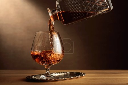 Foto de El brandy se vierte de un decantador en un vaso de francotirador. Coñac sobre una mesa de roble. - Imagen libre de derechos