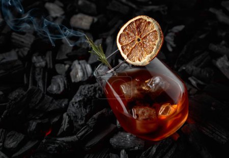 Foto de Cóctel a la antigua con hielo, rebanada de naranja seca y romero. Whisky con romero y hermosos remolinos de humo sobre un fondo de carbón quemado. - Imagen libre de derechos