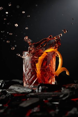 Téléchargez les photos : Un morceau de glace tombe dans le verre avec un cocktail. Cocktail avec glace et écorce d'orange sur charbon de bois brûlant. - en image libre de droit