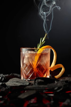 Foto de Cóctel a la antigua con hielo, cáscara de naranja y romero. Whisky con ramita ardiente de romero sobre fondo oscuro. - Imagen libre de derechos