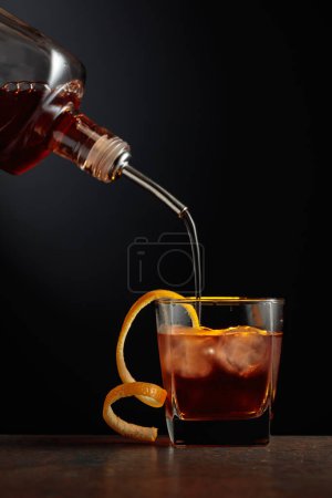 Foto de Cóctel a la antigua con hielo y piel de naranja. Whisky se vierte en un vaso con hielo. - Imagen libre de derechos