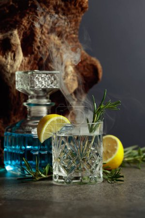 Photo for Cocktail Gin-tonic with lemon and rosemary in a crystal glass. Smoked rosemary old-fashioned cocktail on a table. In the background old snag. - Royalty Free Image