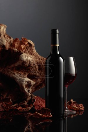 Photo for Bottle and glass of red wine. In the background old driftwood and dried-up vine leaves. Frontal view with space for your text. - Royalty Free Image