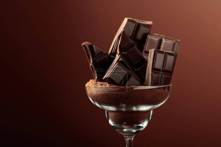 Téléchargez les photos : Barre de chocolat noir cassée en verre avec poudre de cacao. Chocolat sur fond brun avec espace de copie. - en image libre de droit