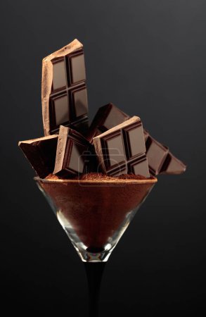 Foto de Barra de chocolate negro roto en vidrio con cacao en polvo. Chocolate sobre fondo negro. - Imagen libre de derechos