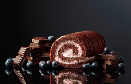 Foto de Pastel de rollo de chocolate con arándanos y una barra de chocolate negra rota sobre un fondo reflectante negro. Copiar espacio. - Imagen libre de derechos