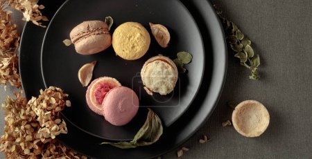 Téléchargez les photos : Délicieux macarons colorés sucrés avec des fleurs séchées sur une assiette noire. Vue du dessus. - en image libre de droit