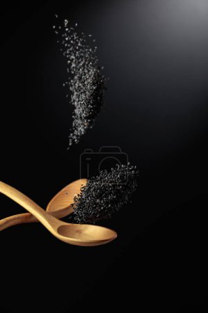 Foto de Los granos de sésamo negro se vierten con cucharas de madera. Sésamo negro sobre fondo oscuro. Copiar espacio. - Imagen libre de derechos
