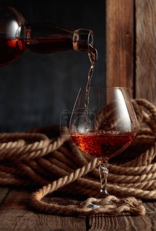 Photo for Rum, cognac, or whiskey is poured into a snifter. Old wooden background with hemp rope. - Royalty Free Image