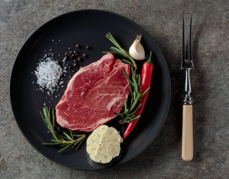 Photo for Raw beef steak for grilling with rosemary, garlic, salt, and pepper. Old stone background. Top view. - Royalty Free Image