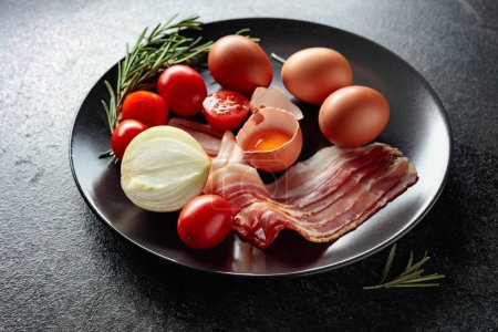 Téléchargez les photos : Bacon fumé avec œufs, tomates, oignons et romarin sur une assiette noire. - en image libre de droit