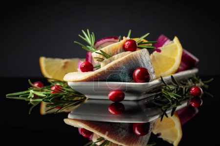 Photo for Herring fillet in oil with lemon, red onion, and rosemary garnished with cranberries. White dish with herring on a black reflective background. - Royalty Free Image