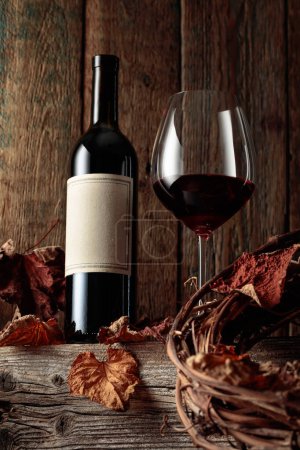 Foto de Botella y copa de vino tinto sobre una vieja mesa de madera con hojas de vid secas. En una botella vieja etiqueta vacía. - Imagen libre de derechos