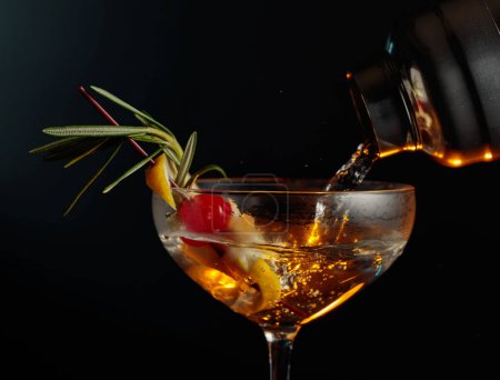 Photo for Martini with cherry, rosemary, and lemon. The drink is poured from a shaker into a glass. - Royalty Free Image