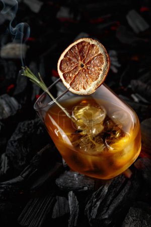 Téléchargez les photos : Whiskey with ice, dried orange slice, and burning rosemary. Whiskey with rosemary and beautiful swirls of smoke on a background of burning charcoal. - en image libre de droit