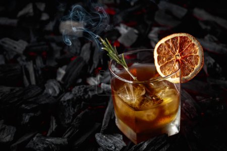 Photo for Whiskey with ice, dried orange slice, and rosemary. Whiskey with rosemary and beautiful swirls of smoke on a background of burning charcoal. - Royalty Free Image
