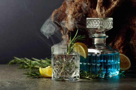 Foto de Cóctel Gin-tonic con limón y romero en una copa de cristal. El romero ahumado el coctel a la antigua sobre la mesa. En el fondo viejo enganche. - Imagen libre de derechos