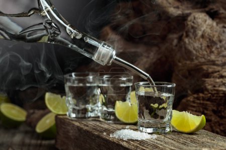 Photo for Tequila is poured from a bottle into a glass. A strong alcoholic drink with salt and lime slices on an old wooden board. - Royalty Free Image