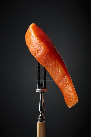 Téléchargez les photos : Smocked salmon on a fork. Fish piece on a black background. - en image libre de droit