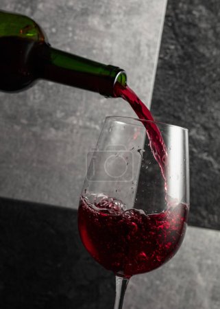 Photo for Red wine is poured from the bottle into a glass. Grey and black stone background. - Royalty Free Image