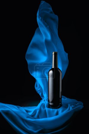 Téléchargez les photos : Bottle of red wine and flutters of blue cloth on a black background. - en image libre de droit