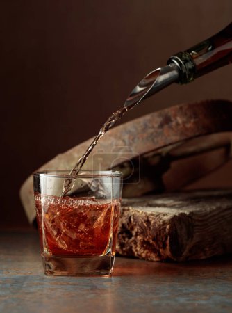 Téléchargez les photos : Le whisky est versé d'une bouteille dans un verre congelé avec de la glace naturelle. Rude fond rouillé. - en image libre de droit