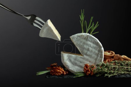 Foto de Queso brie con nueces, tomillo y romero sobre fondo negro. Queso Brie y tenedor con pieza de triángulo. - Imagen libre de derechos