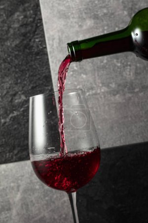 Téléchargez les photos : Le vin rouge est versé de la bouteille dans un verre. Fond en pierre grise et noire. - en image libre de droit