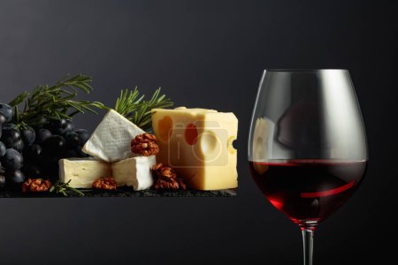 Téléchargez les photos : Vin rouge avec fromage, noix, raisins et romarin sur fond noir. - en image libre de droit