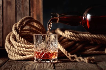 Téléchargez les photos : Le rhum, le cognac ou le whisky sont versés dans un verre de cristal. Vieux fond en bois avec corde de chanvre. - en image libre de droit