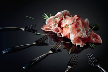Photo for Italian prosciutto or Spanish jamon with rosemary. Meat and forks on a black reflective background. - Royalty Free Image
