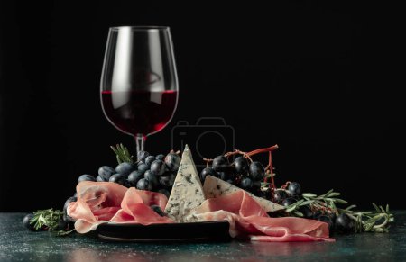 Foto de Vino tinto con uvas, romero, jamón y queso azul sobre un fondo oscuro. - Imagen libre de derechos