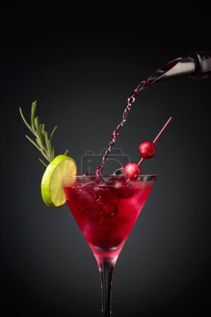 Téléchargez les photos : Cranberry cocktail garnished with berries, lime, and rosemary. In a glass with ice is pouring cranberry liquor. - en image libre de droit