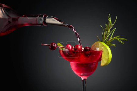 Photo for Cranberry cocktail garnished with berries, lime, and rosemary. In a glass with ice is pouring cranberry liquor. - Royalty Free Image